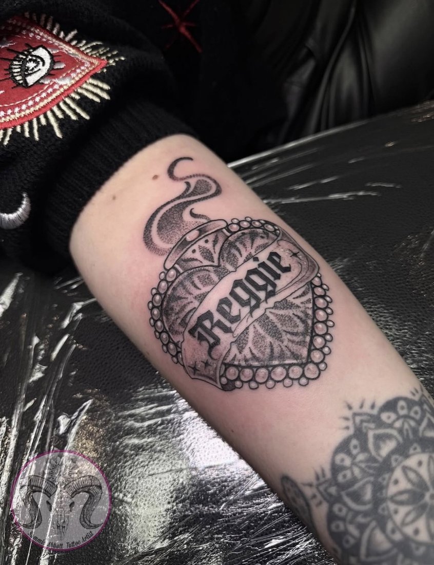 Dotwork locket heart with a banner reading Reggie on the arm